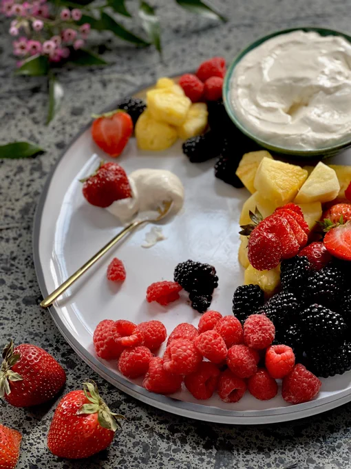 Berries and pineapple on a white plate with a bowl of creamy white fruit dip, and a little gold spoon with some of the dip on a plate.