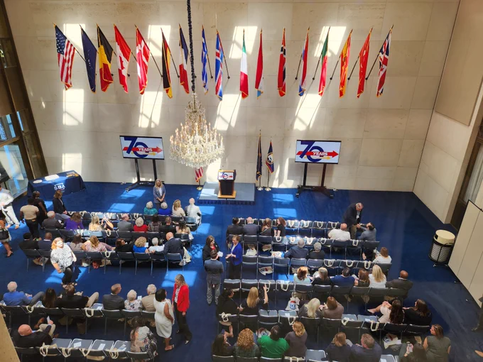 A stage at the Eisenhower Library with flags and a podium flanked by screens showing the SBA 70 logo. A crowd gathers in the seats.