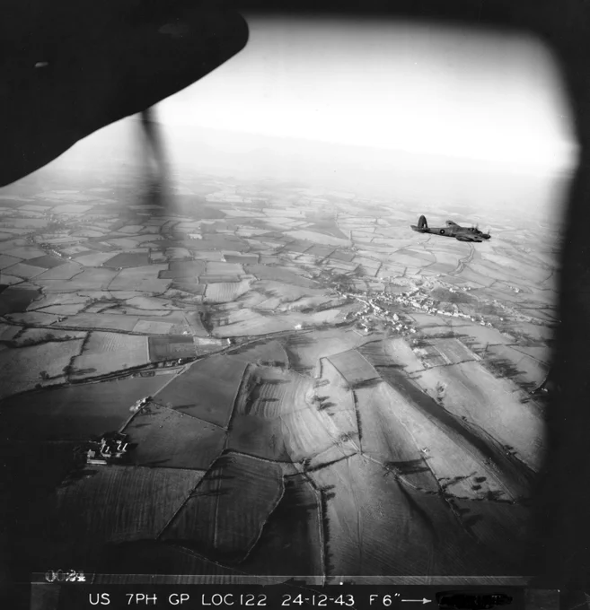 A black and white photograph of a plane flying over farmland.