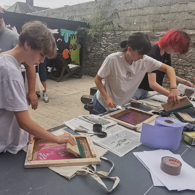 A group of young people are making screen prints on a table in an outside space.