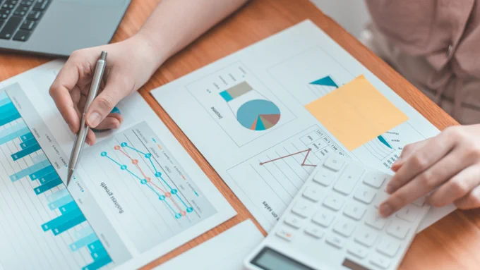 Someone working at a desk using a calculator with print-outs of various data charts in front of them.