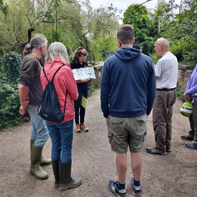 A group of seven people standing on a gravel path with trees in the background. One person in a black polo shirt is presenting from an ID sheet and the others are watching in a semicircle.