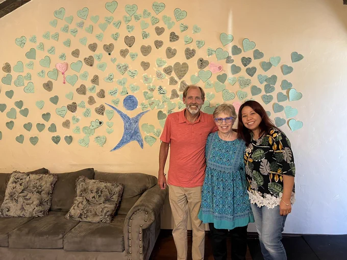 Picture of Jason, Deb and Bel. They are in front of a wall with hearts and an image of a person. They are standing besides a couch. This picture was taken in the living room at SHARE!s peer respite. They are smiling and are happy.