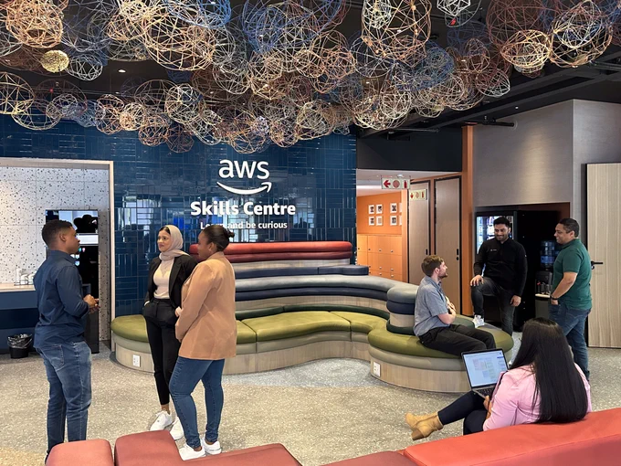Individuals congregating on the main floor of the AWS Skills Center in Cape Town, South Africa. Colorful couch seating lines the walls, and an orb installation lines the ceiling. The wall contains an AWS logo, followed by "Skills Centre" and "Learn and be curious." 