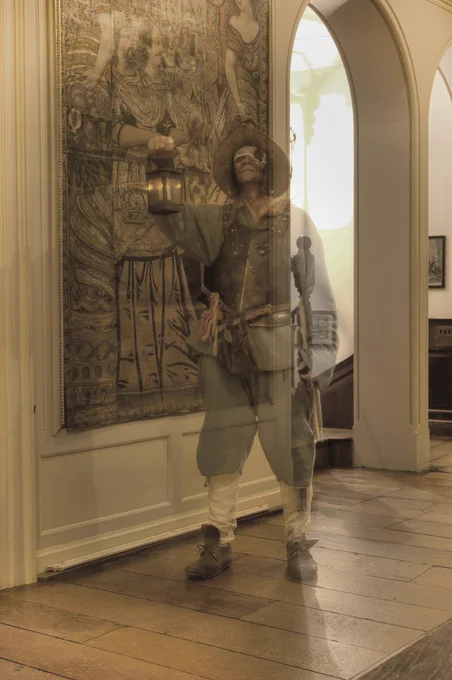 A semi-transparent figure of a man in English Civil War uniform walks past a leather hanging in the Leather Gallery at Dunster Castle. He is holding a lantern and has a bloody bandage over one eye.