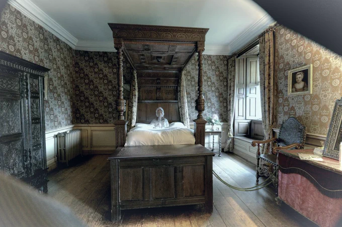 A ghostly grey lady sits in a large four poster bed, she wears a mop cap and night dress and is semi-transparent. 