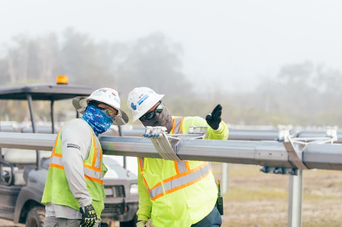 Two individuals in hard hats and high visibility vest installing solar trackers. Making sure the tackers are inline.