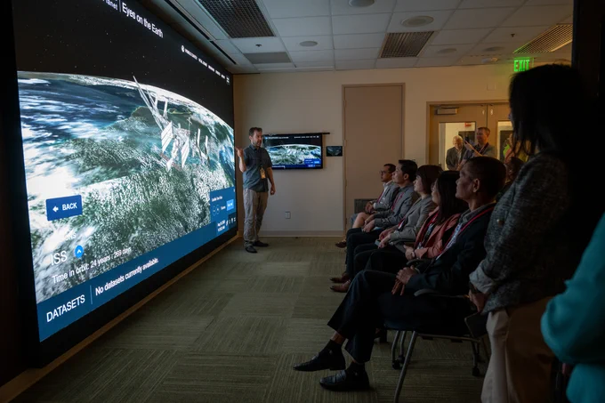 A group of individuals are seated in a small room in front of a giant floor-to-ceiling screen. A presenter stands in front of the screen, pointing to data. On the screen are the words "Eyes on the Earth" with a large image of the Earth. Floating above Earth is a model of the International Space Station. 