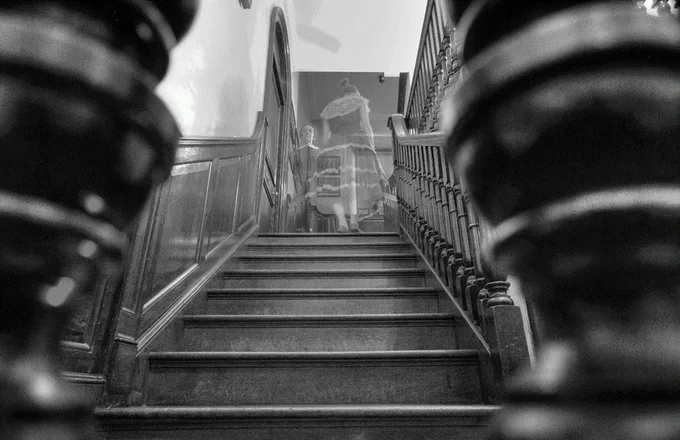 A black and white photograph taken between two bannisters up a staircase, where a young girl in a Victorian dress and a woman in a governess type outfit. They are both semi-transparent, as if they are ghosts 