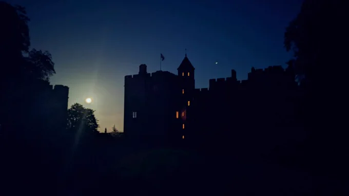 A photograph of Dunster Castle at night. The castle is silhouetted against the night sky with a bright moon to the left. There are orange lights glowing in the windows of a tower in the castle to the left. 