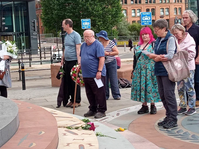 The reading of the names at the Peterloo Memorial at the 2022 commemoration.  A group of people carrying wreaths and flowers, and holding cards with the names of the dead.
