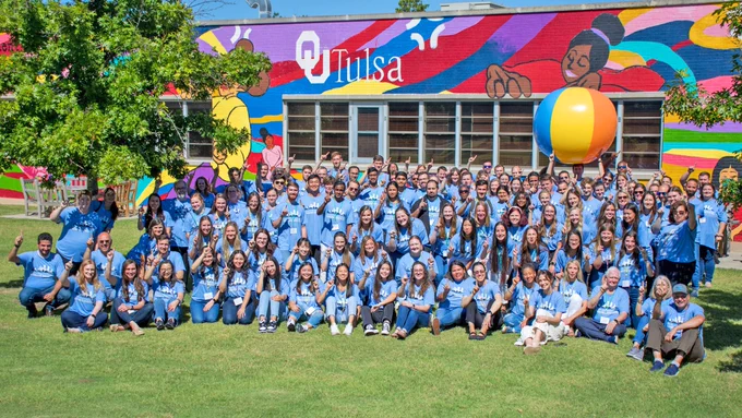 A large group of people in blue shirts sitting in the grass with a brightly colored mural behind them.