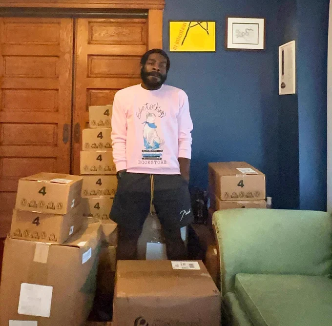 Hanif stands in his living room surrounded by boxes of books and bookbags, wearing a pink sweatshirt and black shorts. The sweatshirt is from Bluestockings Bookstore, and has a blue cat on it, sitting on a pile of books, the top one reading All Cats Are Beautiful.