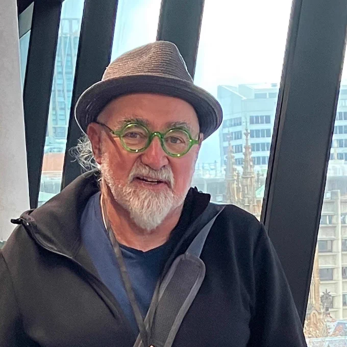 Selected ArtScreen 2023 artist Guy Morgan has a short white beard and green glasses. He is wearing a grey hat and jacket.