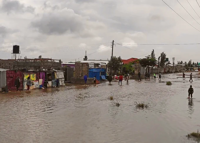 Flooding in Lusaka, Zambia in March 2022. People, infrastructure and trees visible. Grey/ white clouds overhead.