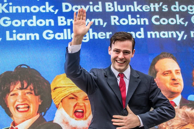 The actor playing Tony Blair is standing by a digi van, he is waving.