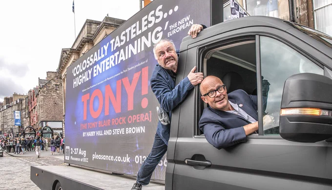 Harry Hill inside a large digivan on the Royal Mile. The van has a large screen which says 'Tony! The Tony Blair Rock Opera!' on it and the quote 'Colossally Tasteless... Highly Entertaining'