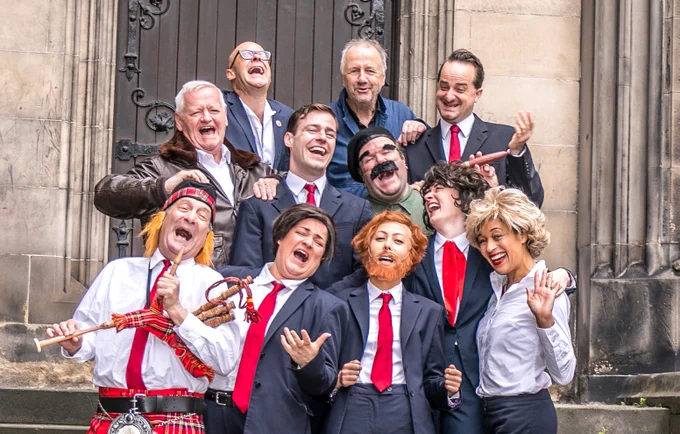 The Tony Blair cast and Harry Hill on the steps of an old building. Their costumes are suits with red ties and they are all laughing.
