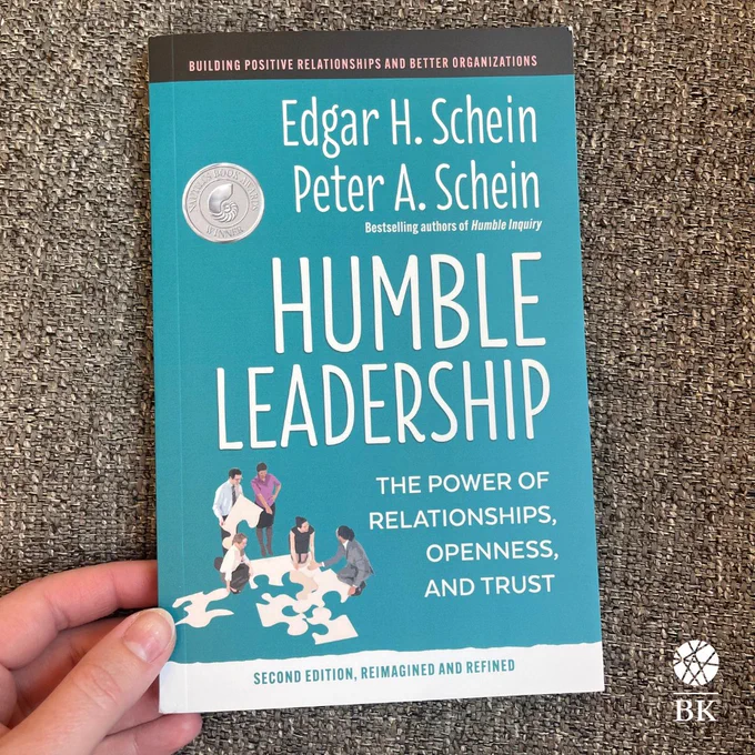 A hand holds a copy of Humble Leadership, Second Edition over a gray fabric.