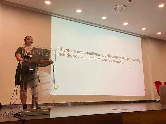 Picture of Dr. Kaitlyn Jones standing on stage at the university of Lusofana giving her keynote presentation to open the second day of the foundations of digital games conference. The current PowerPoint slide on screen shows the joe gerstadt quote “if you do not intentionally, deliberately, and proactively include, you will unintentionally exclude”