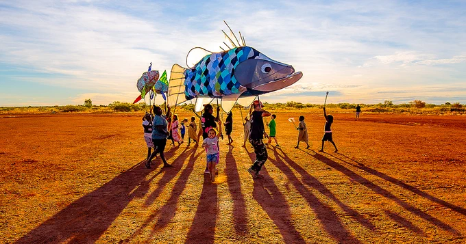 Children holding a large puppet of a fish in the desert with the sun behind it creating interesting shadows.