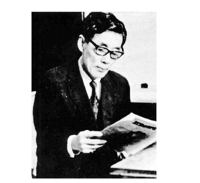 A black and white photograph of Kim Sung hee. He is wearing a dark suit and thick rimmed glasses. He is sat at a desk where he is reading a newspaper.