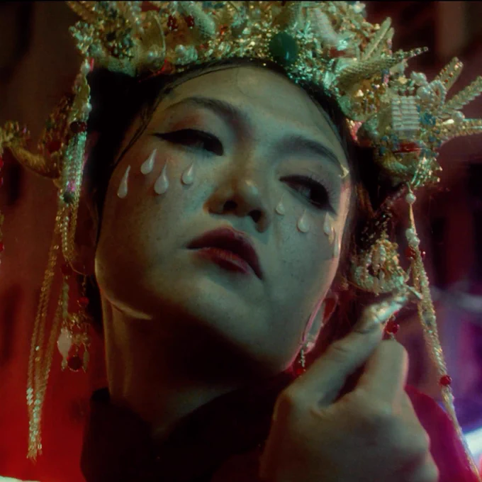 A woman wearing a head dress and makeup to resemble tears looks right of frame