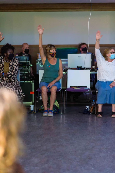 Three women raise one arm in the air as they perform seated dance exercises. The technical team for the event are behind them. All are wearing masks.