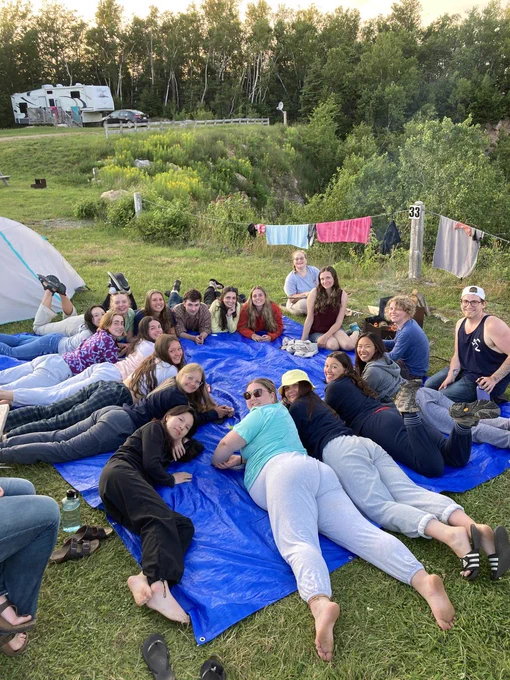 Multiple people laying in a circle, on a blue tarp in a campground.