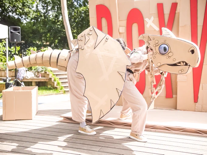 In the centre of the image there is a dragon made from different pieces of cardboard. There is one person with their head inside the head of it and one person holding the wings with the tail attached at the back. They are stood close together to create the look of a whole dragon.