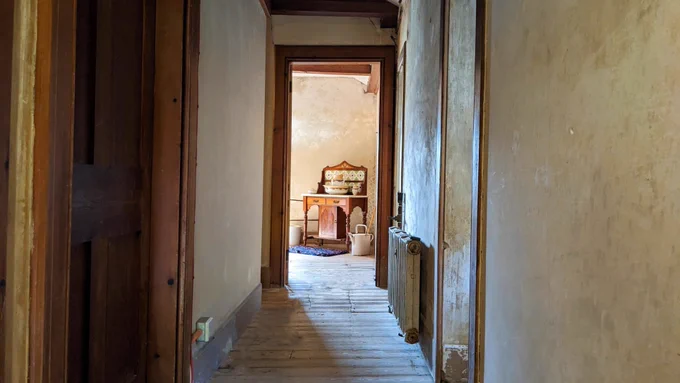 A photograph down a corridor towards and open door with a wooden wash stand, pitcher, bucket and rug visible through the doorway 