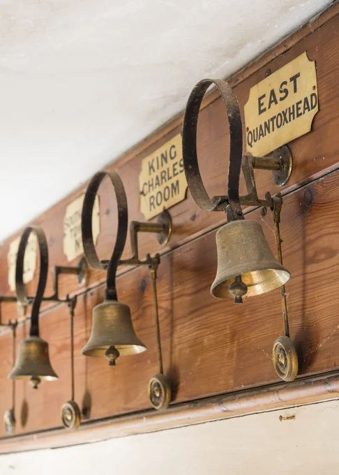 A row of bells mounted on a wooden board. There are room labels on the wall, the closest reads "East Quantoxhead" and the next reads "King Charles Room", the rest of the labels are illegible. 