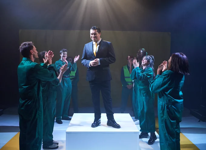A production shot of an actor wearing a grey suit and a yellow tie standing on a white platform. A group of people circle around whilst clapping.
