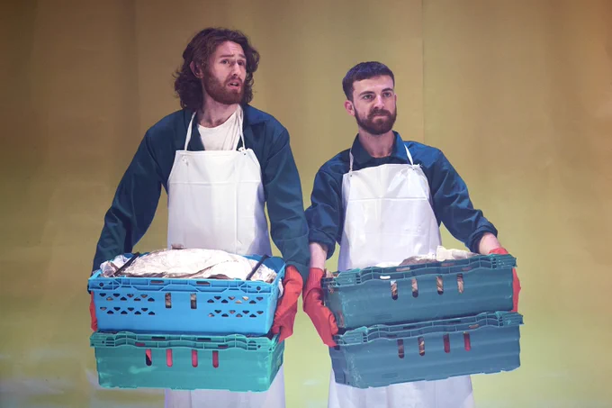 A production shot of two actors standing next to each other carrying green grocery baskets. They wear green boiler suits, a white apron and red gloves.