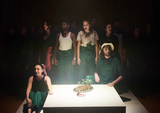 A production shot of a group of actors wearing green boiler suits. Most of them stand in a line and two of them kneel down by a white platform. On the platform are a set of seedlings in pots and a photograph.