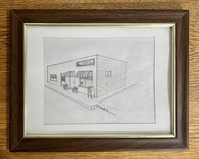 A framed pencil perspective drawing of a building with awnings over a door and a window, planters, and a sign that reads "Basehor Community Library."