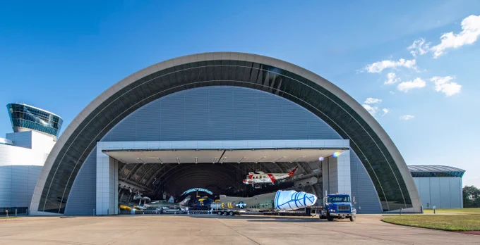 B-17 bomber with its nose wrapped in a covering, is towed by a blue semi-truck and backed into a hangar at the Steven F. Udvar-Hazy Center.