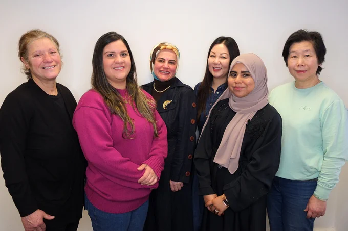 A group of 6 women from Shelanu Women's Craft Collective stand against a white wall smiling at the camera