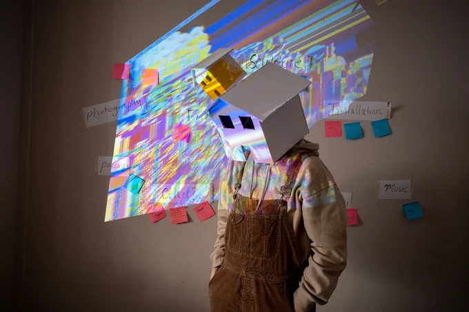 A person wearing dungarees stands with a mask shaped like a house on their head. A colourful light projection distorts across the mask