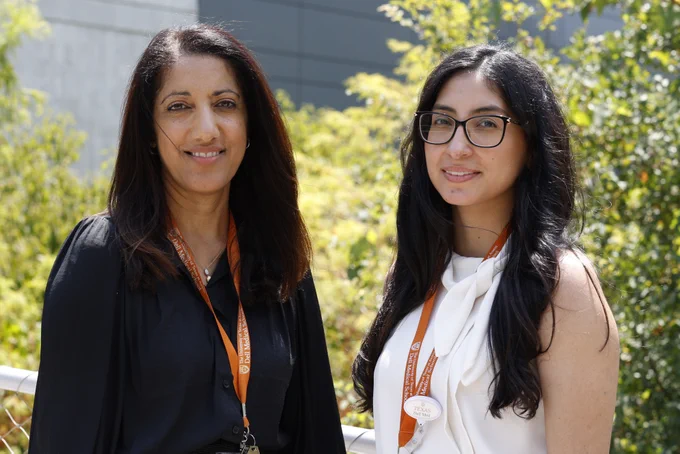 Image of Professor Kiran Shokar and Research Program Manager Cynthia Chacon standing in front of Dell Medical School. They are both wearing UT Dell Medical School lanyards and are smiling towards the camera. Photo by Connor Steen.