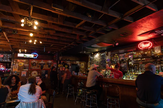  Image shows the inside of Hole in the Wall. The image shows a diagonal angle of the bar. There are people sitting at the bar as well as tables enjoying drinks. The lighting is dim and grungy with lots of stickers on the wall as well as a few neon signs. Image by Lorianne Willett.
