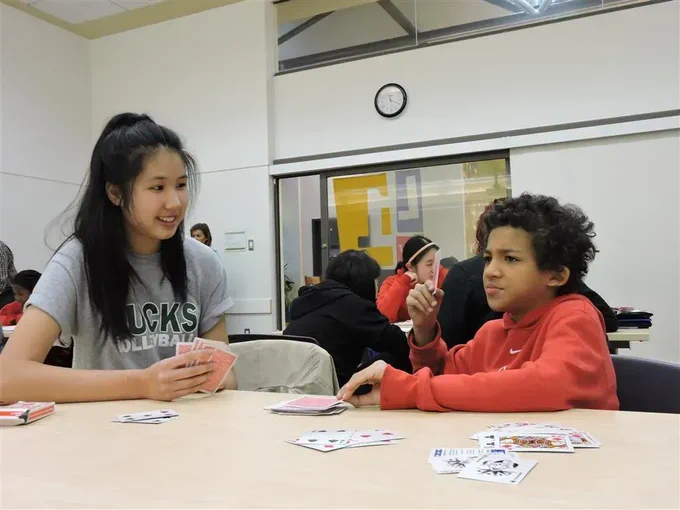 Two kids sitting at a table playing cards in a room full of other kids doing similar activities.