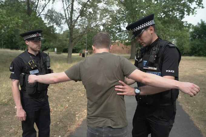 Two uniformed Essex Police officers speaking to a man in a T-shirt who is standing with his arms outstretched. One officer is searching the man, the other is watching