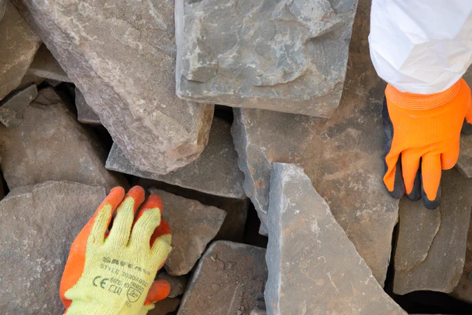 A collection of angular looking grey rocks with two gloved hands, as if the people are selecting a stone from a pile.