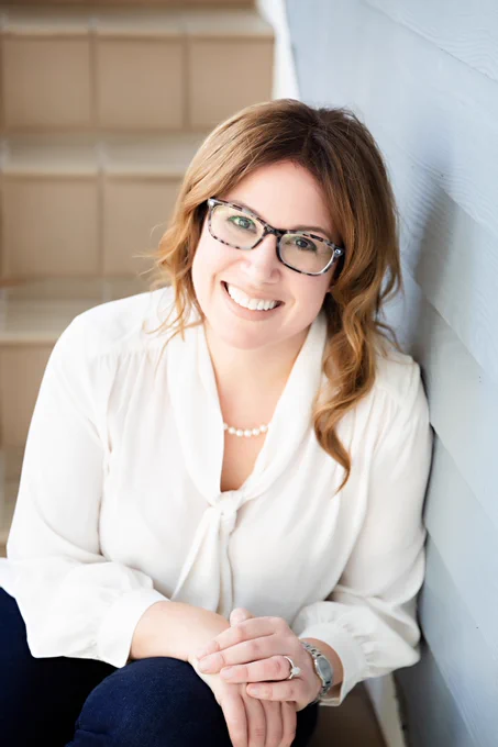 A portrait image of Stacey Steinberg leaning against a white wall with tan steps in the background.