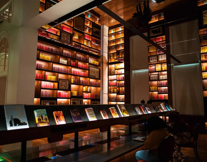 The magic Library at Planet Word. There are books in shades of yellow and red going up to the ceiling, which is mirrored to make the bookshelves look twice as tall. In the center of the room are tables with books on display above the table surface.