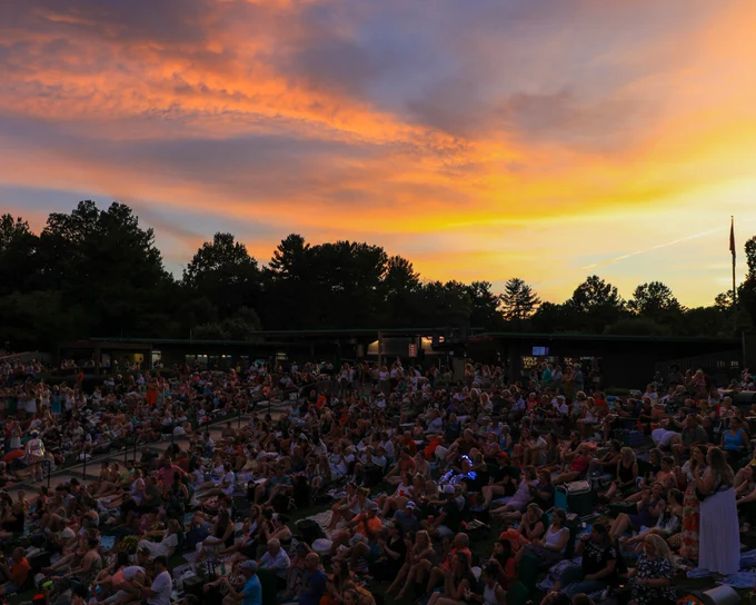 A large crowd of people sit on a lawn with a yellow-orange sunset in the background.