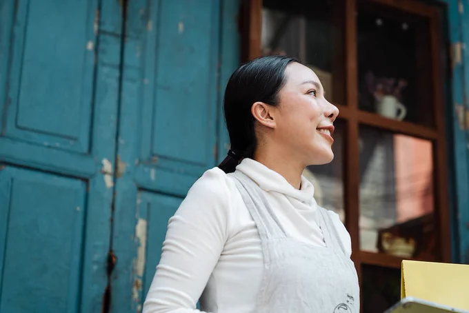 Happy smiling Asian female in apron standing against blue shabby weathered building and looking away dreamily