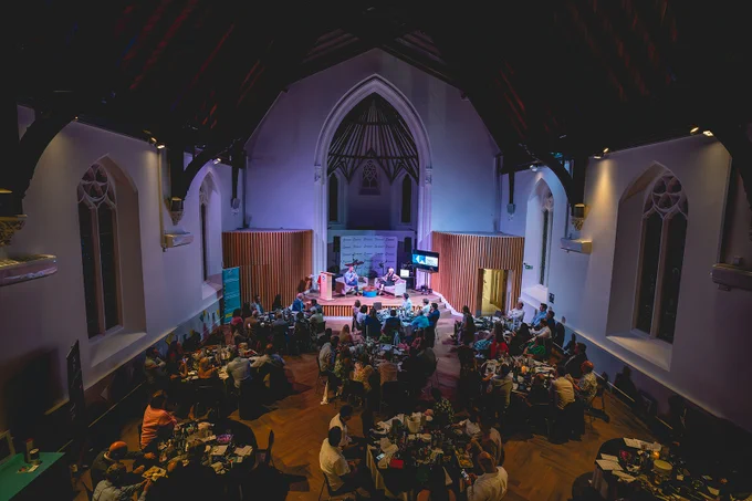 Image of a church hall. There are nine round tables, filled with people sitting at them. At the very front, is a small stage with two people sat on chairs, chatting.