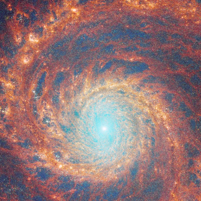 A large spiral galaxy takes up the entirety of the image. The core is mostly bright white, but there are also swirling, detailed structures that resemble water circling a drain. There is white and pale blue light that emanates from stars and dust at the core’s centre, but it is tightly limited to the core. The rings feature colours of deep red and orange and highlight filaments of dust around cavernous black bubbles.